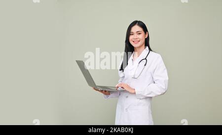 Professional and gorgeous Asian female doctor in white gown and stethoscope holding a opened laptop, standing against grey studio background. Stock Photo