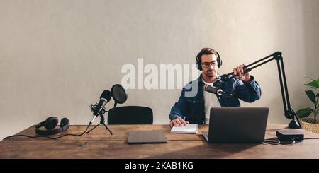 Radio dj adjusting his microphone while sitting behind a laptop with headsets. Male podcaster preparing to go live on air in a home studio. Young cont Stock Photo