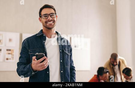 Smiling businessman with eyeglasses holding a smartphone while standing in an office. Happy young businessman working in a creative and modern workpla Stock Photo