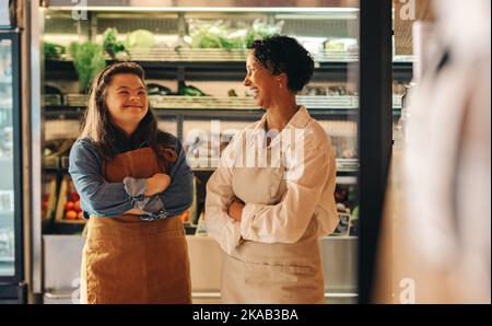 Two grocery store workers laughing cheerfully while standing together in their supermarket. Happy woman with Down syndrome running a small business wi Stock Photo