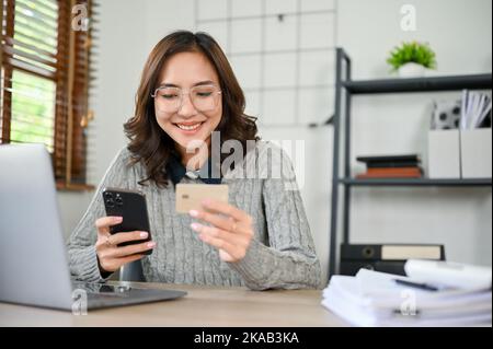 Beautiful millennial Asian businesswoman or female office worker in glasses sitting at her desk, holding her smartphone and credit card. online paymen Stock Photo