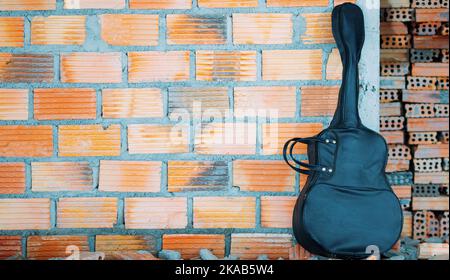 Inside the house - black acoustic guitar case, brown brick wall background. Stock Photo