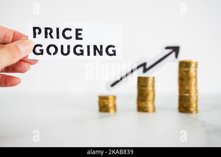 hand holding Price Gouging text in front of growing stacks of coins in the background, symbol of excessive pricing or speculation Stock Photo