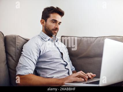 Working from home is always the best. a focused young man working on his laptop while being seated on a couch at home during the day. Stock Photo