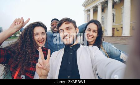 Point of view shot of multiracial group of tourists taking selfie in city center holding gadget camera and posing together with hand gestures expressing positive emotions. Stock Photo