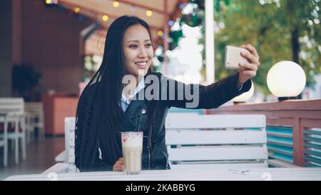 Happy Asian lady is talking to friends online making video call using smartphone sitting in open-air cafe alone. Girl is talking, touching long black hair and smiling. Stock Photo
