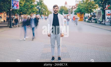 Portrait of young businessman in trendy garments standing in busy pedestrian street and looking at camera while crowds of people are whizzing around. Stock Photo