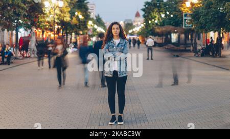 Portrait of stylish young lady tired of usual haste standing in the street among whizzing people and looking at camera. Time, youth and modern society concept. Stock Photo