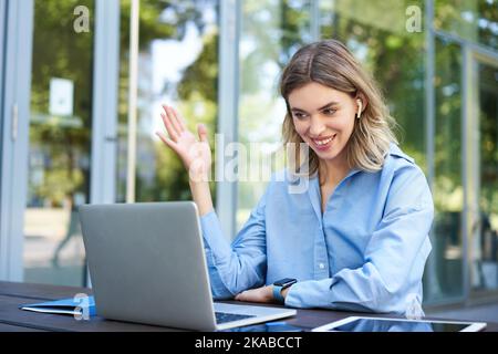 Portrait of corporate woman working outside office on remote, sitting with laptop, wireless earphones, attend online video conference Stock Photo