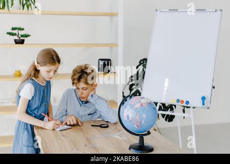 Teenage girl and boy siting near table with globe in classroom. Teen schoolboy and schoolgirl doing project together at school Stock Photo