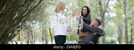 Two joyful woman and a man in wheelchair give high five with their hands Stock Photo
