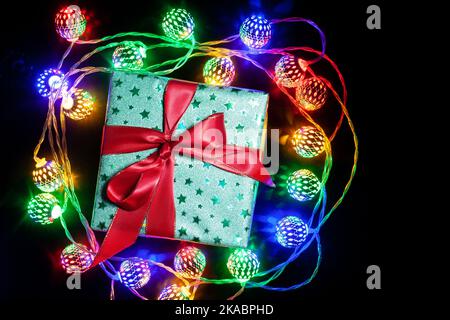 Single green gift box with red ribbon bow on black surface with multicolor garland lamps. Gift box wrapped in green shine paper with red ribbon. Top v Stock Photo