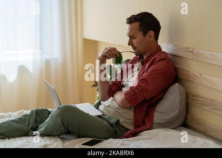 Young man freelancer sits on bed in hotel room with laptop on lap keeping track of new job orders  Stock Photo