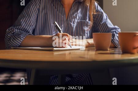 Female hand with pen taking notes, writing plans in diary on wood table with coffee cups in coffeehouse. Woman at wooden desk with planner and tea mug Stock Photo