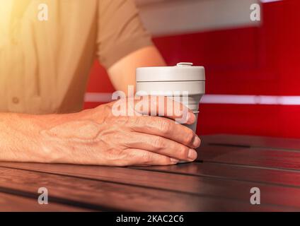 Male hands closeup holding reusable silicon takeaway eco coffee cup in hands over wooden street table on court. Stock Photo