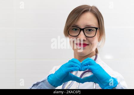Beautiful young happy doctor woman in medical gloves and glasses with a stethoscope in the hospital shows hand gestures. Selective focus. Portrait. Cl Stock Photo