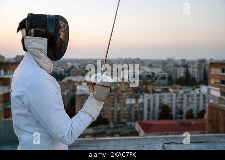 Portrait of young woman fencer wearing mask, white fencing costume and practicing outdoors. Stock Photo