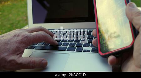 Hands of a man using a laptop and smartphone. He is sitting outside. Home office. Mobile working Stock Photo
