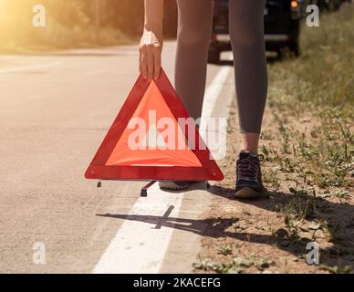 Young women hands close up putting triangle caution sign on road near broken car on roadside. Stock Photo