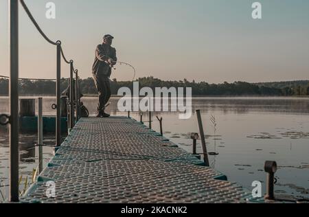 Fisherman fishing in foggy early morning. Man with flying fish rod resting on river at dawn. Stock Photo