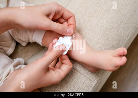 Mother woman sticks a medical adhesive plaster on the toddler baby leg. Mom s hand with sticky wound protection tape and child s foot. Kid aged one ye Stock Photo