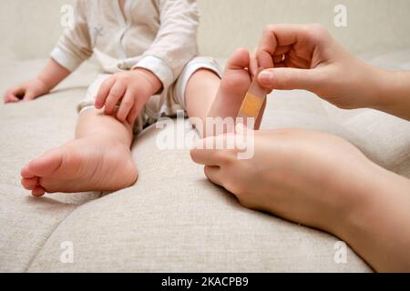 Mother woman sticks a medical band-aid on the toddler baby leg. Mom s hand with sticky wound protection tape and child s foot. Kid aged one year and t Stock Photo