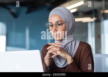Close-up photo of satisfied business woman looking at laptop screen, muslim woman in hijab working inside modern office building, female boss thinking about decision. Stock Photo