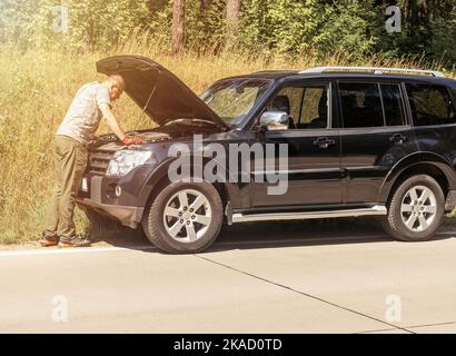 Driver standing near car hood raised up and checking, inspecting for problems of broken auto on road side. Stock Photo