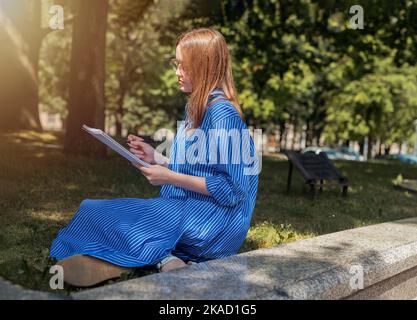 Young female student sitting on grass with notepad oracademic book and learning, Reading and writing in park. Stock Photo