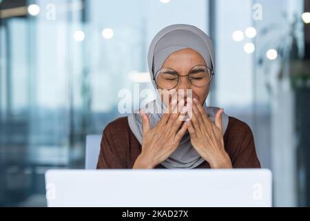 Close-up photo of businesswoman in hijab tired working with laptop yawning sitting on chair inside modern office building. Stock Photo