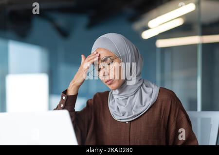 Sick woman in hijab working inside modern office building, businesswoman in glasses has severe headache, close-up photo with laptop. Stock Photo