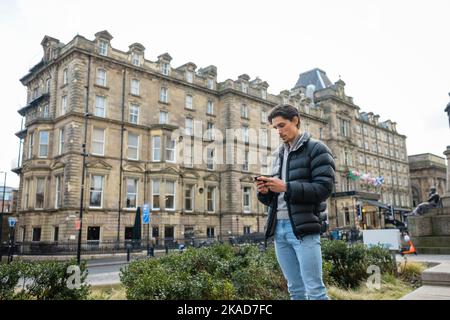 A wide-view shot of a young man standing in the city centre, he is using his smartphone to send a text message. Stock Photo