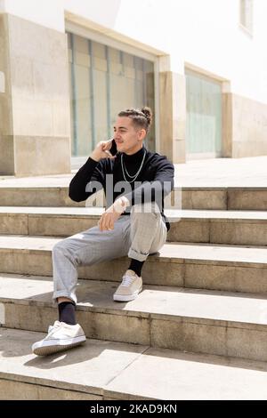 Portrait of a smiling young mixed-race man talking on a smartphone sitting on stairs in the city. communication, mobile apps, business, lifestyle Stock Photo