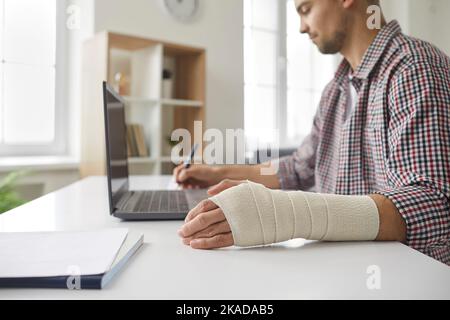 Man with elastic bandage on his hand sitting at table, working on laptop and writing in notebook Stock Photo
