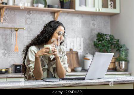 Beautiful hispanic woman with curly hair at home sitting in kitchen and watching video content on laptop, woman holding cup of hot drink in hands smiling. Stock Photo