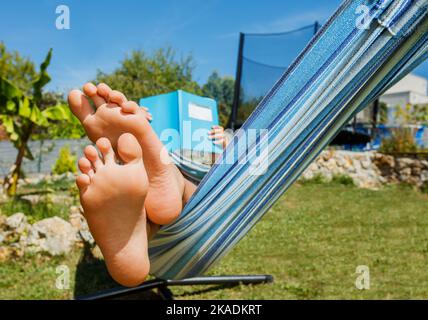 Boy rest in hammock reading a book, feet on foreground Stock Photo
