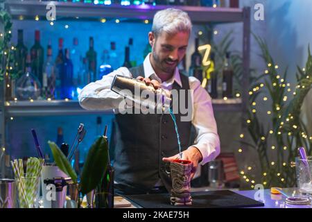 Professional barman in a white shirt and black apron making blue cocktail pouring alcohol at party in nightclub. Nightlife. Stock Photo