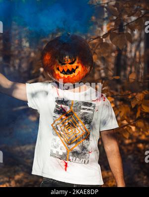 A man wearing pumpkin on his head in a park during fall with blue smoke in the background Stock Photo