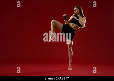 Female trainer showing kick exercise for self defence by legs on red background. Front view of attractive woman training in black sport bandages in studio. Concept of box. Stock Photo
