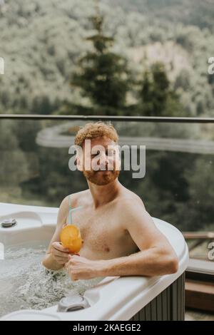 Attractive young man enjoying in outdoor hot tub and drink fresh orange juice on vacation Stock Photo