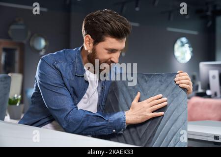 Bearded man in denim shirt exploring soft pattern chair before buying on shopping mall. Professional male designer checking texture of furniture textile, while touching chair back. Concept of design. Stock Photo