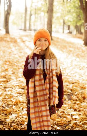 Portrait of smiling school aged girl isolated on sunny yellow forest background. Happy child in warm cozy hat, scarf and sweater walk in golden park Stock Photo