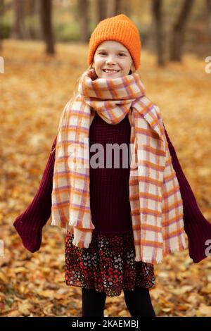 Portrait of happy school aged girl in cute warm hat, scarf and cozy knitted sweater isolated on dry yellow fallen foliage background. Autumn activity Stock Photo