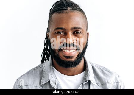 Close-up photo of an attractive black guy, african american man, with dark brown eyes, with dreadlocks, standing over isolated white background, looks at camera with candid pleasant smile Stock Photo