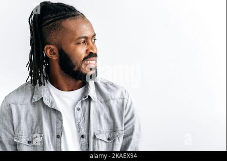 Photo of an attractive black guy, african american man, with dreadlocks, in casual shirt, standing over isolated white background, looking to the side with candid pleasant smile. Copy-space concept Stock Photo