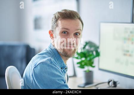 Cute bearded caucasian male freelance software engineer sitting at working place in home office hardly working using computer. Young handsome developer man in denim shirt. High quality image Stock Photo