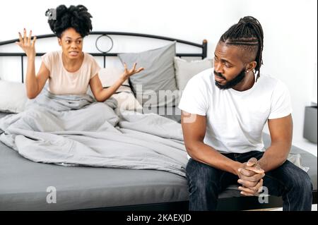 Relationship problems, disagreements. Upset african american husband sits on bed, his wife scolds him, screams emotionally and gesticulates with hands. Quarrel between spouses, conflict Stock Photo