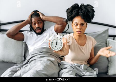 Oversleep concept, being late for work. Shocked stunned african american marriage couple sitting on a bed in bedroom looking in amazement at camera, woman holds alarm clock in hand Stock Photo