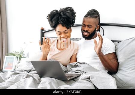 Distant video communication. Positive happy african american spouses, husband and wife, are sitting in a bed in bedroom, using laptop, talking by video conference with friends or family, smiling Stock Photo