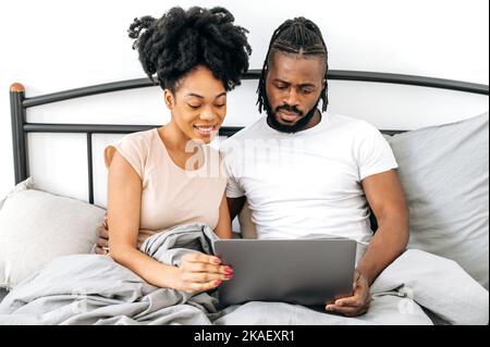 Positive, satisfied african american spouses, husband and wife, are sitting in a bed in bedroom, using laptop, browsing internet, watching movie together before going to bed, reading news, smiling Stock Photo